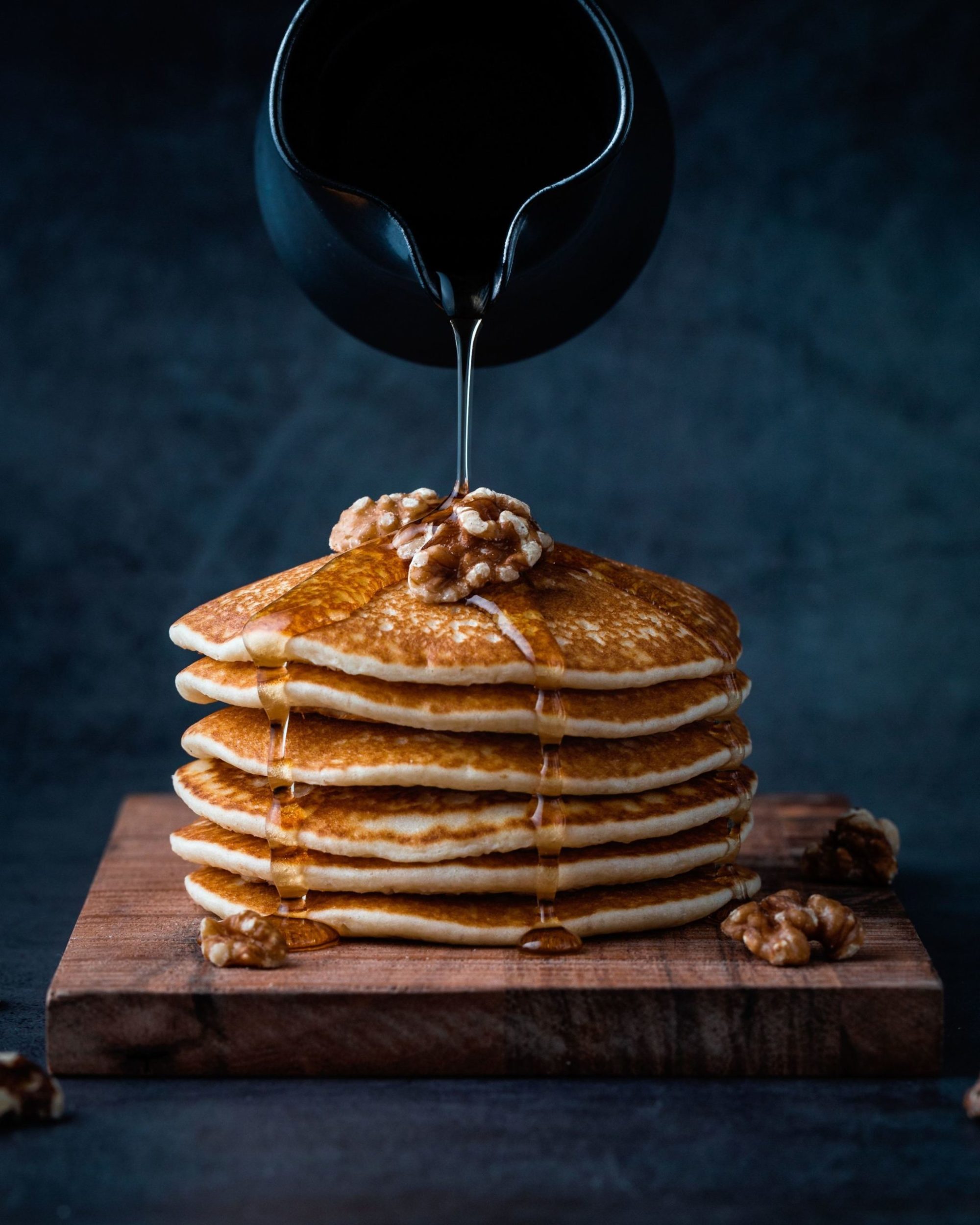 pitcher of maple syrup being poured over a stack of pancakes with walnuts