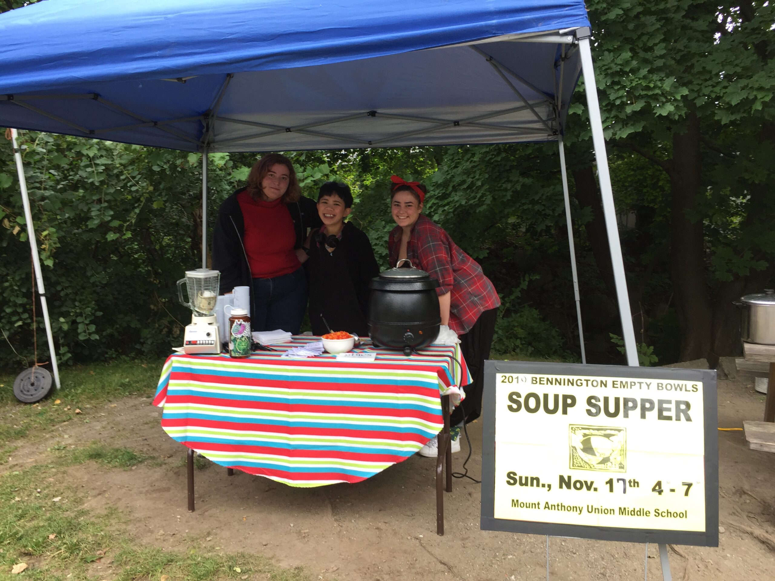 Three young people stand behind a table under a pop-up tent smiling and serving soup with a sign for Bennington Empty Bowls Soup Supper