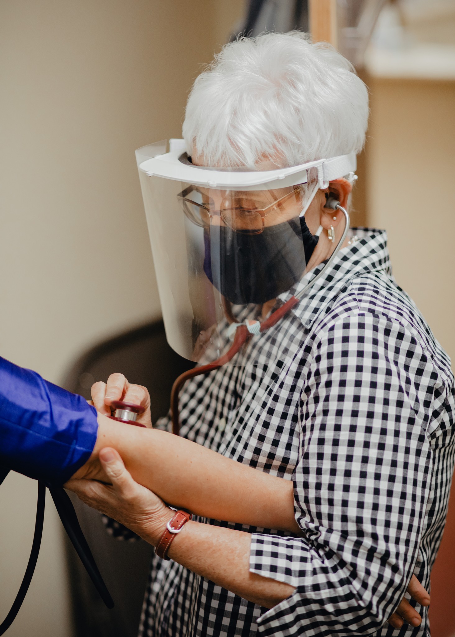 A woman with short white hair, glasses and face mask checks a patient's blood pressure